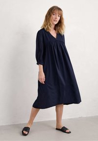 Tamsiai mėlyna, ilgomis rankovėmis suknelė su V formos iškirpte ir susiraukšlėjusia juosmeniu, pagaminta iš lengvos medžiagos; derinama su juodais, perforuotais šliopais.