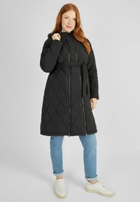Abrigo largo negro acolchado con capucha, presenta una cintura ceñida con cinturón, dos bolsillos delanteros con cremallera y un acabado suave y brillante. Ajuste y diseño casual.
