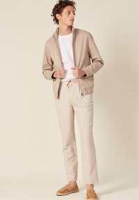 Beige leichte Jacke, weißes T-Shirt, hellbeige Hose und braune Slipper. Geradliniges Design mit minimalen Details und weichen Texturen.