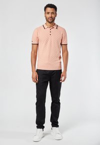 Vaaleanpunainen polo-paita, jossa on teksturoitu kuvio, mustia yksityiskohtia kauluksessa ja hihoissa; yhdistetty mustiin housuihin ja valkoisiin tennareihin.