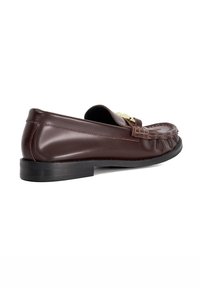 Brown leather loafer with a smooth finish, textured stitching, and a matte black outsole. Features a gold-toned accent on the vamp.
