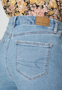 Světlé modré džínové kalhoty s vysokým pasem, s koženou štítkem označeným "AMERICAN EAGLE" a standardními zadními kapsami s jemným prošíváním.