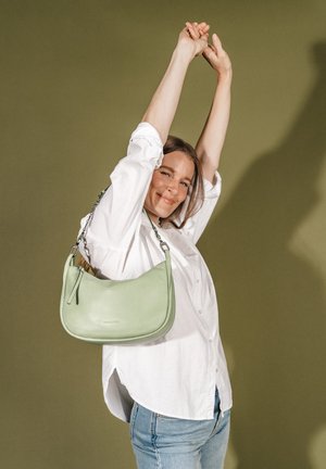 Frau in weißem Hemd und Jeans streckt die Arme über den Kopf, lächelt, mit einer hellgrünen Schultertasche vor einem schlichten olivgrünen Hintergrund.