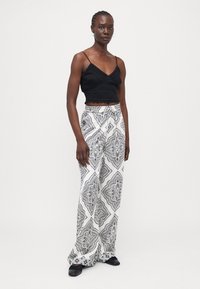 Top negro corto con textura de encaje; pantalones blancos de pierna ancha con patrones geométricos negros intrincados; modelo con zapatos negros.
