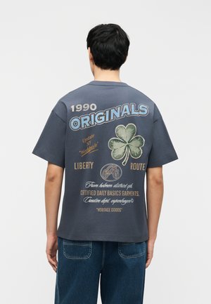 Homme de dos portant un T-shirt gris foncé avec le texte "1990 ORIGINALS", un grand motif de trèfle vert et un texte imprimé supplémentaire au dos.