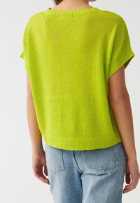Žena oblečená v jasně zeleném pleteném topu s krátkým rukávem, doplněná o světle modré džíny, zobrazená zezadu na jednobarevném pozadí.