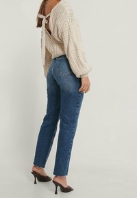 Mujer vestida con un suéter de punto crema con lazo en la parte de atrás, jeans azul de cintura alta y mules marrones con tacón, de pie contra un fondo ligero y sencillo.