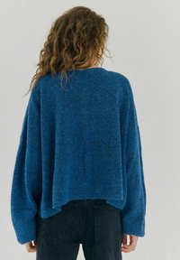 Maglione blu lavorato a maglia con una vestibilità ampia, maniche larghe e schiena corta. Texture morbida con un motivo melangiato. Indossata sotto ci sono jeans neri.