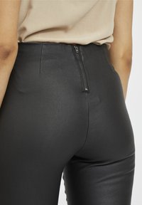 Personne portant un pantalon en cuir noir taille haute avec une fermeture éclair à l'arrière et un haut beige rentré à l'intérieur, vue de la taille vers le bas.
