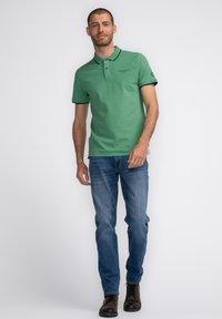 Zelené polo tričko s kontrastním tmavým límečkem a knoflíky, v kombinaci s modrými džíny a hnědými botami. Jednoduchý design, bavlněný materiál.