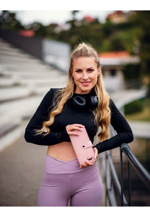 Pink wallet with a snap closure and polka dot accent, held alongside black headphones and worn with a black long-sleeve top and lavender leggings.