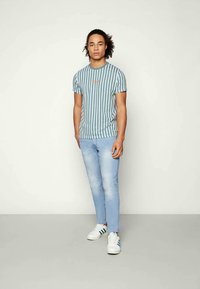 Τιρκουάζ και λευκή ριγέ κοντομάνικη μπλούζα, συνδυασμένη με ανοιχτό μπλε τζιν και λευκά αθλητικά παπούτσια με πράσινες λεπτομέρειες. Απλό φόντο.