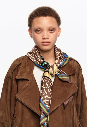 Joven con cabello corto y rizado, que lleva una chaqueta de ante marrón y una bufanda de seda marrón con estampado, anudada al cuello.