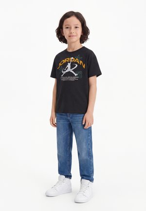 Enfant debout avec un léger sourire, portant un t-shirt noir Jordan, un jean bleu et des baskets blanches sur un fond blanc uni.