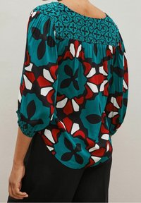 Blusa colorata con un motivo floreale nei toni del verde teal, rosso e nero. La parte superiore ha un collo arrotondato e maniche a sbuffo di lunghezza 3/4.