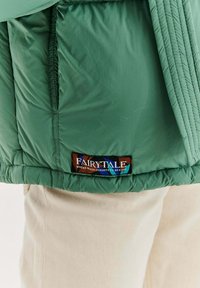 Zielony dół kurtki z wielokolorową metką z napisem „Bajka, w której magia zapomina być miła”, noszony na beżowych spodniach.