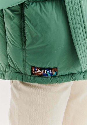 Zielony dół kurtki z wielokolorową metką z napisem „Bajka, w której magia zapomina być miła”, noszony na beżowych spodniach.