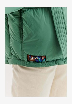 Zielony dół kurtki z wielokolorową metką z napisem „Bajka, w której magia zapomina być miła”, noszony na beżowych spodniach.