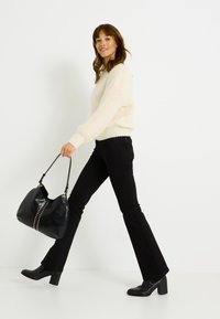 Bolso de cuero negro con tachuelas doradas, llevado por una persona con un suéter crema de peluche y jeans negros acampanados, caminando contra un fondo blanco.