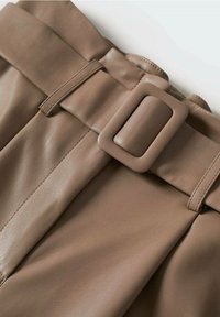 Gros plan sur un pantalon en cuir marron avec une large ceinture assortie et une boucle rectangulaire, sur un fond clair.