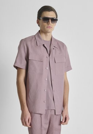 Uomo che indossa una camicia color malva a maniche corte con bottoni e pantaloni abbinati, con occhiali da sole scuri, in piedi davanti a uno sfondo chiaro uniforme.