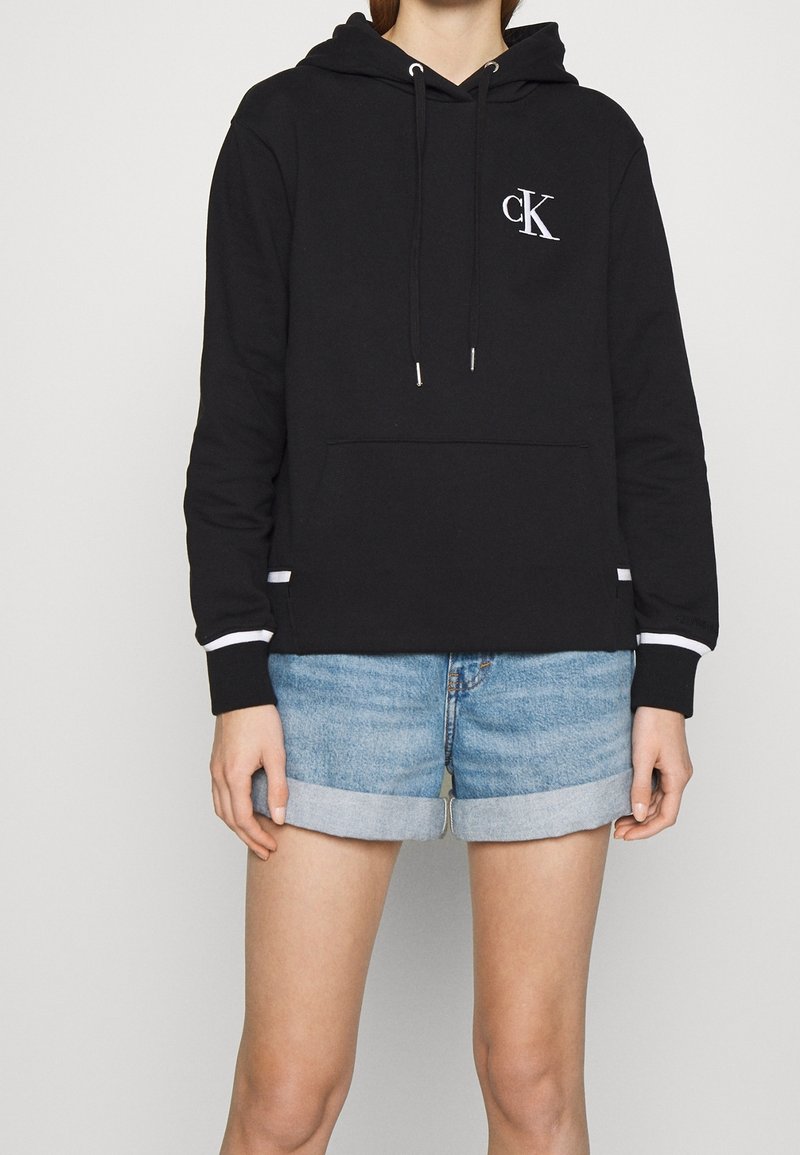 Femme portant un sweat à capuche Calvin Klein noir avec logo blanc et un short en denim retroussé sur un fond uni.