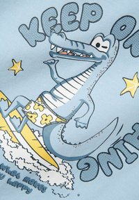 Tela azul claro con un caimán de dibujos animados surfeando, vestido con un bañador de flores. El texto dice "SIGUE ADELANTE" con estrellas adicionales y un diseño de ola.