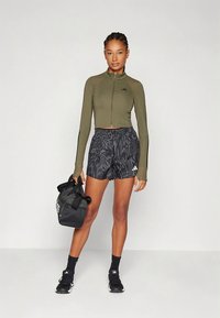 Olivgrüne, taillierte, gekürzte Zip-up-Jacke, schwarze gemusterte Shorts, schwarze Turnschuhe und eine schwarze Sporttasche. Eng anliegendes Design mit sportlichem Stil.