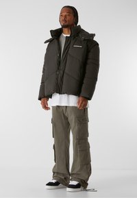 Mattschwarze Pufferjacke mit geometrischer Naht; lockere Passform. Kombiniert mit olivgrünen Cargo-Hosen mit mehreren Taschen. Schwarze Sneakers.