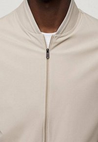 Hellbeige Bomberjacke mit Stehkragen, Reißverschluss vorne und glatter Textur. Verfügt über ein minimalistisches Design und weiches Gewebe.