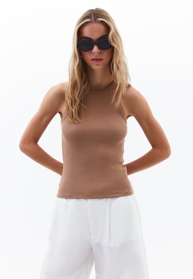 Top senza maniche di colore beige a costine con collo alto, abbinato a pantaloni bianchi a gamba larga. Il modello indossa occhiali da sole neri. Design minimalista, texture liscia.
