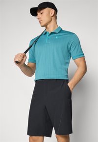 Turkizna polo majica s golim ovratnikom i tamnim akcentima, uparena s crnim kratkim hlačama. Model drži golf palicu. Nošena crna kapa.