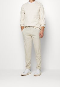 Beige sweatshirt med en framficka, tillsammans med ljusbeige byxor. Vita sneakers fulländar outfiten. Slät textur genom hela plagget.