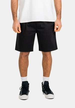 Mann trägt schwarze knielange Shorts, weiße Sneakersocken, schwarze Turnschuhe und ein weißes Hemd, steht mit entspannt an den Seiten hängenden Armen.