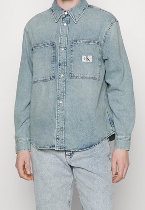 Hellblaue Jeansjacke mit zwei Brusttaschen, silbernen Druckknöpfen und einem kleinen Calvin Klein Logo-Patch auf der rechten Tasche, getragen mit passenden Jeans.