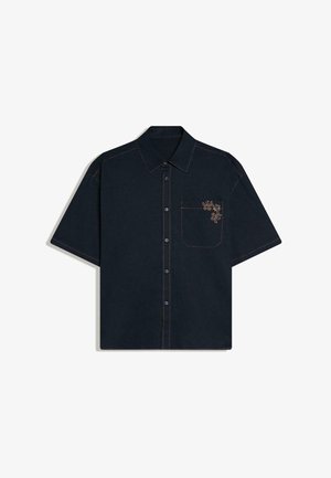 Črna srajca s kratkimi rokavi in gumbi, z ozkim ovratnikom, sprednjim žepom na levi strani prsnega koša ter bež cvetličnim vezenjem na vogalu žepa.