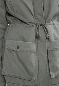 Olivgrüne Jacke mit Kordelzug in der Taille, ausgestattet mit zwei aufgesetzten Vorderaschen und einem glatten, strukturierten Stoff.