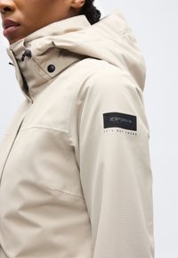 Gros plan d'une personne portant une veste imperméable beige avec capuche et un écusson noir « Icepeak » sur le bras gauche au niveau du haut de la manche.