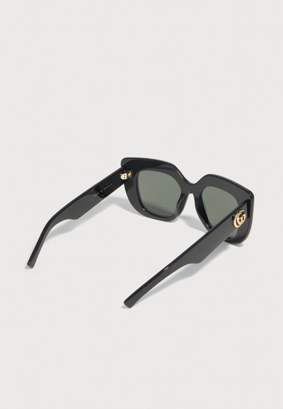 Lunettes de soleil carrées noires oversize avec verres teintés foncés et ornements logo dorés sur les branches, présentées sur un fond clair.