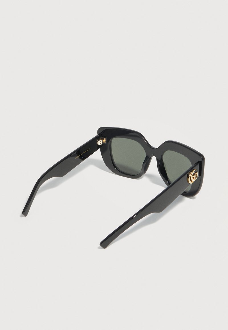 Occhiali da sole quadrati oversize neri con lenti scure e decorazioni con logo dorato sulle aste, esposti su uno sfondo chiaro.