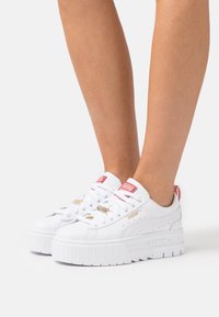 Białe trampki na platformie z gładkiej skóry, z złotymi i czerwonymi akcentami oraz grubą, teksturowaną gumową podeszwą. Sznurowadła są białe z złotymi elementami.