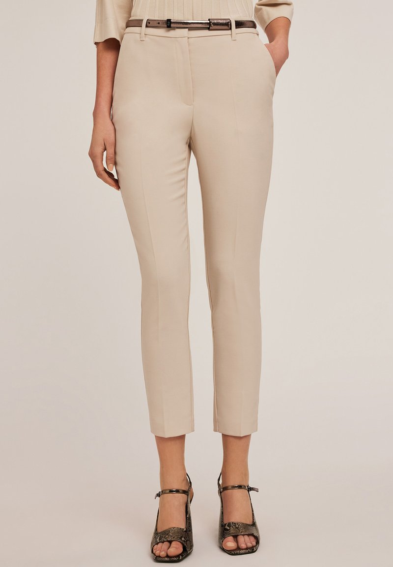 Pantaloni beige su misura con vestibilità slim, dotati di cintura e tessuto liscio. Lunghezza cropped con pieghe stirate, abbinati a tacchi texturizzati.