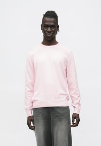 Ανοιχτό ροζ φούτερ με στρογγυλή λαιμόκοψη, ριμπ μανσέτες και τελείωμα. Φέρει μικρό λογότυπο στο στήθος. Φορώντας το με σκούρο γκρι τζιν παντελόνι.