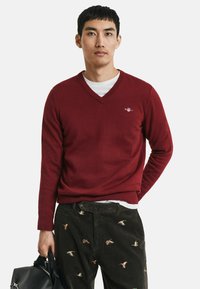 Burgunderfarbener V-Ausschnitt-Pullover aus weichem Stoff, mit langen Ärmeln und gerippten Bündchen, über einem weißen Hemd getragen, kombiniert mit detaillierten dunklen Cordhosen.