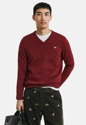 Burgunderfarbener V-Ausschnitt-Pullover aus weichem Stoff, mit langen Ärmeln und gerippten Bündchen, über einem weißen Hemd getragen, kombiniert mit detaillierten dunklen Cordhosen.