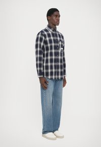Marine- og hvit rutete flanellskjorte med knapper, enkelt brystlomme; brukt med lyseblå jeans og hvite sneakers. Enkelt bakgrunn.