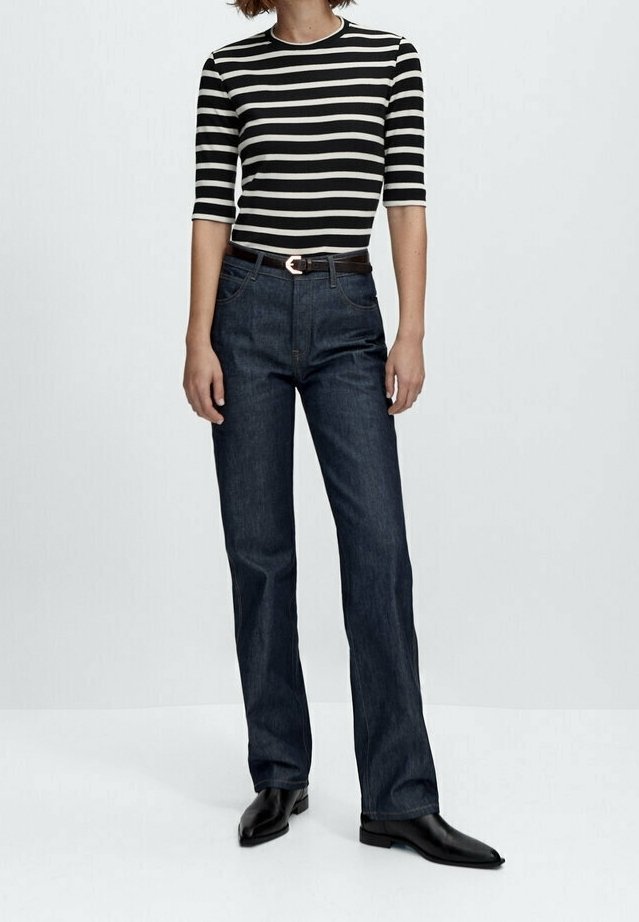Personne portant une chemise rayée noire et blanche, un jean bleu foncé droit avec ceinture, et des chaussures noires à bout pointu, debout sur un fond blanc.