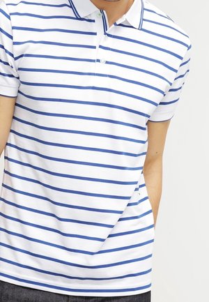 Mann trägt ein weißes Poloshirt mit blauen horizontalen Streifen und steht mit einer Hand in der Tasche.