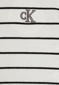 Biela bavlnená tkanina s čiernymi horizontálnymi pruhmi. Obsahuje malé šedé vyšívané logo "cK" na ľavej strane. Mäkšia textúra.