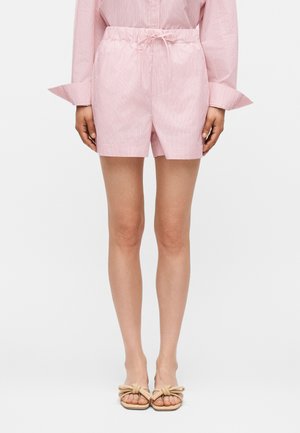 Nő világos rózsaszín csíkos rövidnadrágot visel rugalmas derékpánttal és megkötővel, hozzá illő hosszú ujjú inggel és bézs papuccsal.